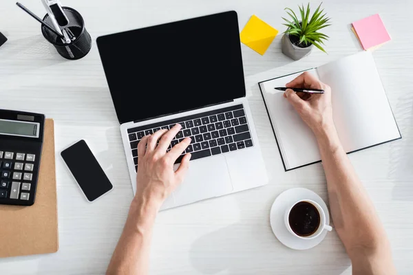 Vue du dessus de l'homme dactylographiant sur l'ordinateur portable et écrivant sur l'ordinateur portable près du téléphone portable et de la papeterie sur la table, gagnant le concept en ligne — Photo de stock