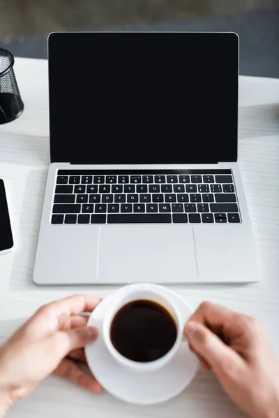 Vista recortada del hombre sosteniendo la taza de café cerca del ordenador portátil con pantalla en blanco, ganando concepto en línea - foto de stock
