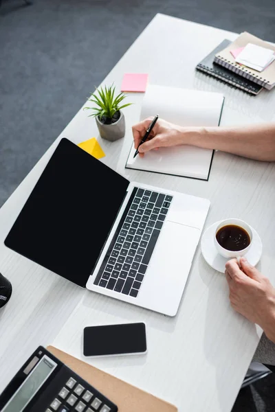 Ausgeschnittene Ansicht eines Mannes, der auf einem Notizbuch in der Nähe von Gadgets auf dem Tisch schreibt und Online-Konzept verdient — Stockfoto