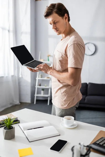 Selective focus of concentrated man using laptop and looking at notebook at home, earning online concept — Stock Photo