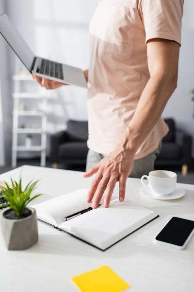 Selective focus of man near notebook with smartphone and plant on table at home, earning online concept — Stock Photo