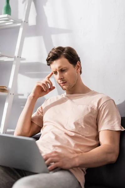 Selective focus of pensive teleworker using laptop on sofa in living room, earning online concept — Stock Photo