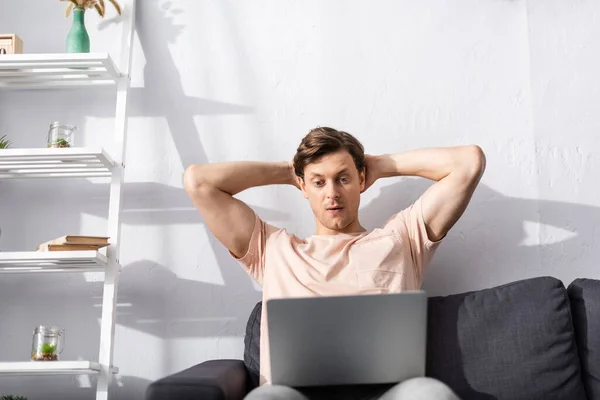 Selective focus of concentrated freelancer with hands behind head looking at laptop on couch, earning online concept — Stock Photo