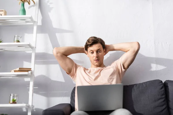 Enfoque selectivo del freelancer concentrado con las manos detrás de la cabeza mirando el portátil en el sofá, ganando concepto en línea - foto de stock