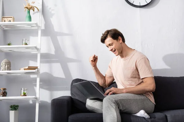 Hombre positivo mostrando sí gesto mientras se utiliza el ordenador portátil en la sala de estar, concepto de ganar en línea - foto de stock