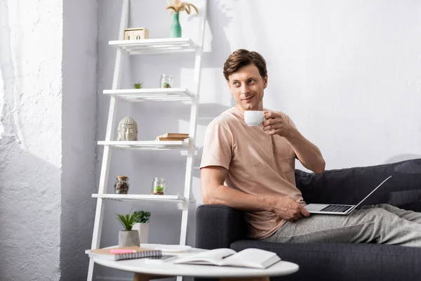 Selective focus of smiling freelancer drinking coffee and using laptop near stationery on coffee table, earning online concept — Stock Photo