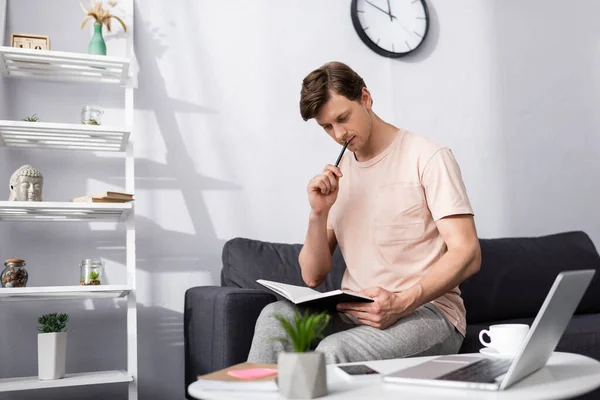 Selective focus of pensive freelancer holding notebook and pen near gadgets and coffee on coffee table, concept of earning online — Stock Photo