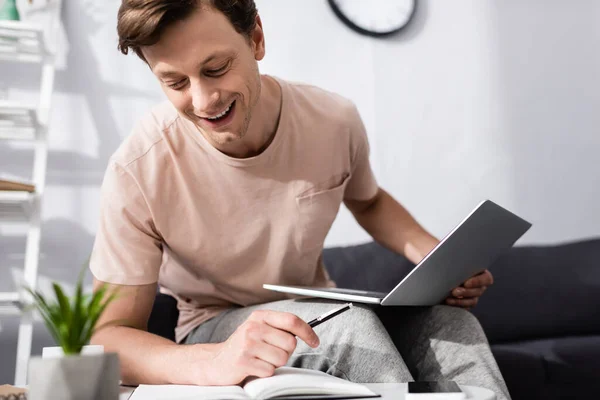 Selektiver Fokus des lächelnden Freiberuflers, der Stift und Laptop in der Hand hält, während er zu Hause das Notizbuch betrachtet, Konzept des Online-Verdienens — Stockfoto