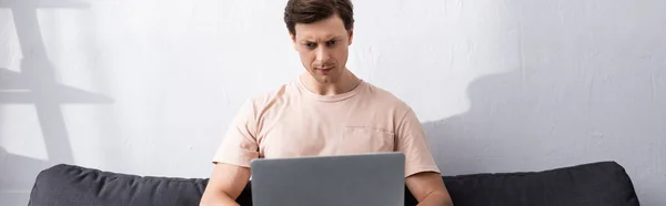 Panoramic shot of concentrated man using laptop on couch at home, earning online concept — Stock Photo