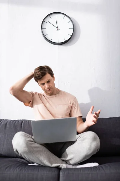 Enfoque selectivo de freelancer reflexivo mirando el ordenador portátil mientras está sentado en el sofá, ganando concepto en línea - foto de stock