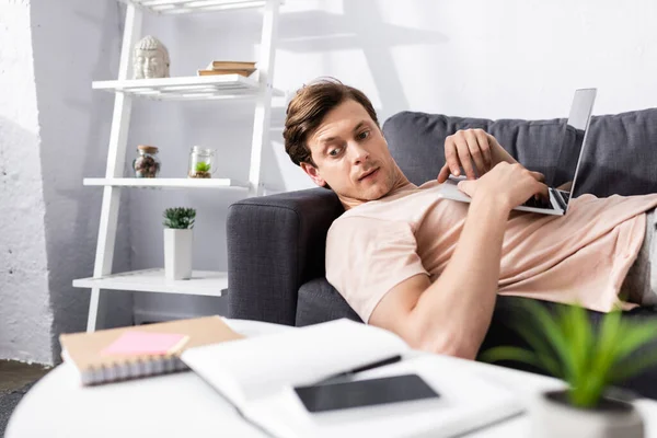 Enfoque selectivo del freelancer que mira el teléfono inteligente y el portátil en la mesa de café mientras usa el portátil en el sofá en casa, concepto de ganar en línea - foto de stock