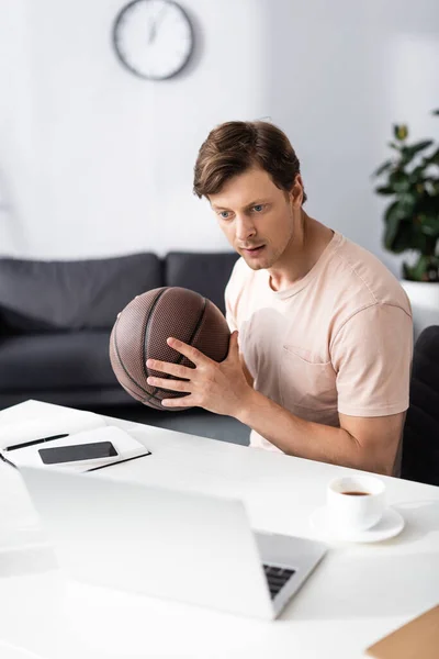 Focus selettivo del bel freelance in possesso di basket e guardando computer portatile vicino alla tazza di caffè sul tavolo, guadagnando concetto online — Foto stock