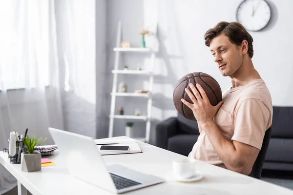 Foco seletivo de homem bonito segurando basquete perto de laptop e papelaria na mesa em casa, conceito de ganhar on-line — Fotografia de Stock