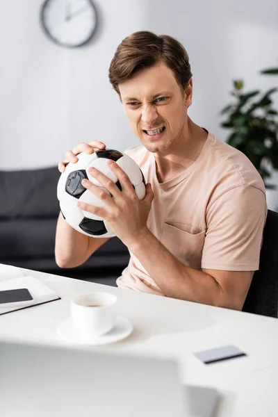 Enfoque selectivo del hombre enojado que sostiene el fútbol cerca de gadgets y tarjeta de crédito en la mesa, concepto de ganar en línea - foto de stock