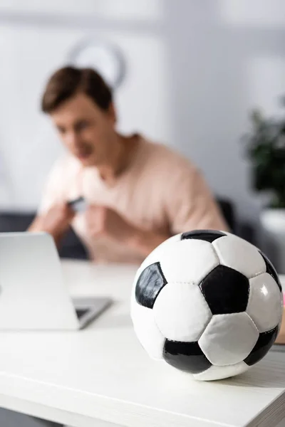 Selektive Fokussierung des Fußballs auf Tisch und Mann, der zu Hause neben Laptop sitzt, Konzept des Online-Verdienens — Stockfoto