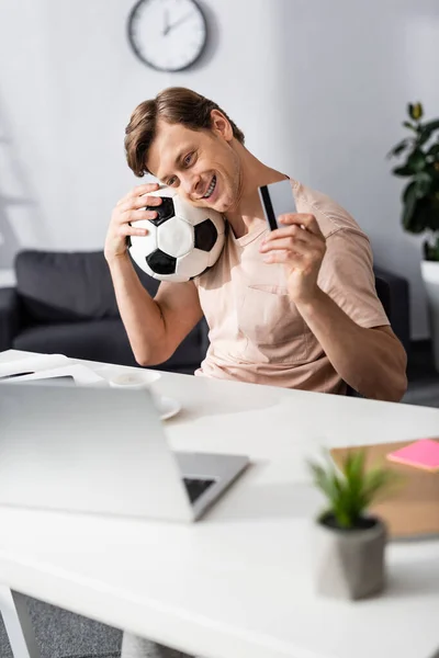 Enfoque selectivo del hombre sonriente que sostiene la tarjeta de crédito y el fútbol cerca de la computadora portátil en la mesa, ganando concepto en línea - foto de stock