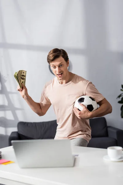 Enfoque selectivo del hombre emocionado sosteniendo efectivo y fútbol cerca de la computadora portátil en la mesa en casa, concepto de ganar en línea - foto de stock
