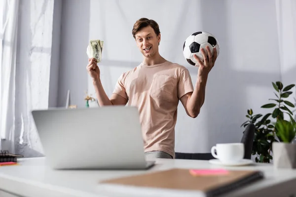 Concentration sélective de l'homme joyeux tenant le football et l'argent près de l'ordinateur portable à la maison, concept de gagner en ligne — Photo de stock