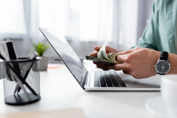 Vista recortada del hombre contando billetes de dólar cerca de la computadora portátil y la taza de café en la mesa, ganando concepto en línea - foto de stock