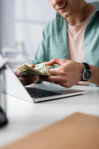 Vista cortada de homem sorridente contando dólares perto de laptop na mesa em casa, ganhando conceito online — Fotografia de Stock