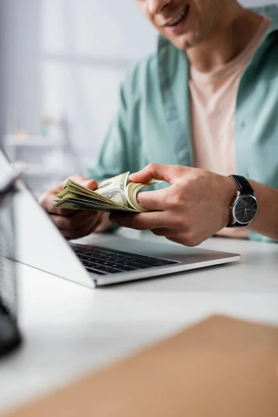 Vista recortada del hombre sonriente contando dólares cerca de la computadora portátil en la mesa en casa, ganando concepto en línea - foto de stock