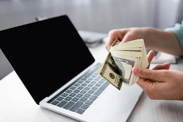 Cropped view of man holding dollar banknotes near laptop with blank screen on table, earning online concept — Stock Photo