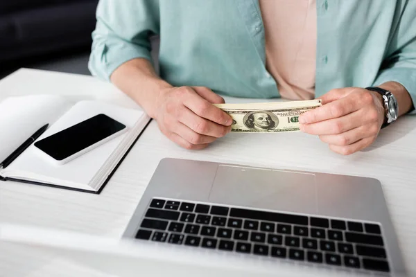 Cropped view of man holding dollars near laptop and smartphone with blank screen on table, concept of earning online — Stock Photo