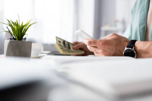 Ausgeschnittene Ansicht eines Mannes mit Smartphone und weißen Dollars am Tisch im Wohnzimmer sitzend, Online-Konzept verdienen — Stockfoto