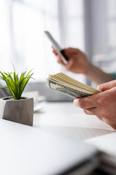 Cropped view of man holding money and using smartphone near plant on table at home, earning online concept — Stock Photo