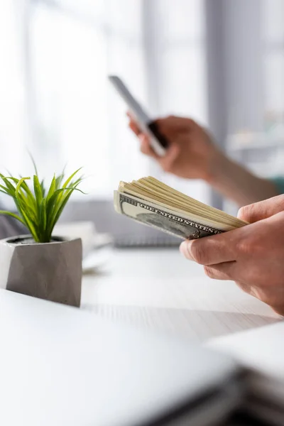 Vista recortada del hombre que sostiene el dinero y el uso de teléfonos inteligentes cerca de la planta en la mesa en casa, ganando concepto en línea - foto de stock