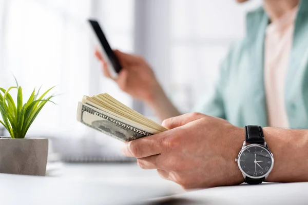 Vista recortada del hombre sosteniendo dólares y utilizando el teléfono inteligente en casa, ganando concepto en línea - foto de stock