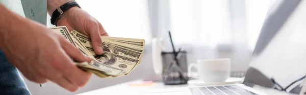 Panoramaaufnahme eines Mannes, der Dollarnoten in der Nähe eines Laptops auf dem heimischen Tisch zählt und sich damit ein Online-Konzept verdient — Stockfoto