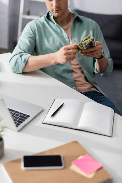 Cropped view of man holding cash near digital devices and notebooks on table at home, earning online concept — Stock Photo