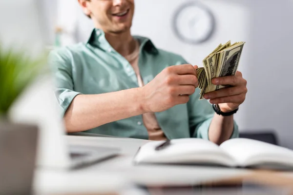Ausgeschnittene Ansicht eines lächelnden Freiberuflers, der Dollars neben Laptop und Notizbuch auf dem Tisch zählt und Online-Konzept verdient — Stockfoto