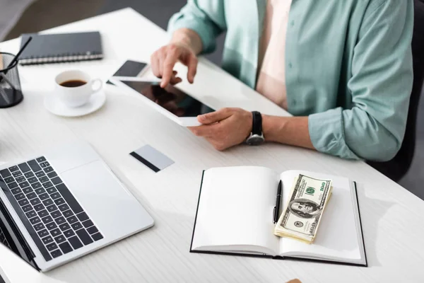 Обрезанный вид человека с помощью цифрового планшета возле гаджетов, кофе и долларов на ноутбуке на столе, зарабатывая онлайн концепция — стоковое фото