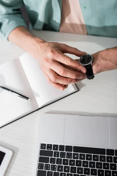 Vista recortada del freelancer verificando el tiempo en el reloj de pulsera cerca del portátil y el portátil en la mesa, concepto de gestión del tiempo - foto de stock