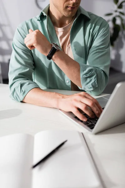 Ausgeschnittene Ansicht eines Mannes mit Laptop in der Nähe eines offenen Notizbuchs mit Stift auf dem Tisch — Stockfoto