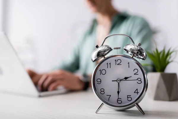 Enfoque selectivo del reloj despertador en la mesa cerca del hombre utilizando el ordenador portátil en casa, concepto de gestión del tiempo - foto de stock