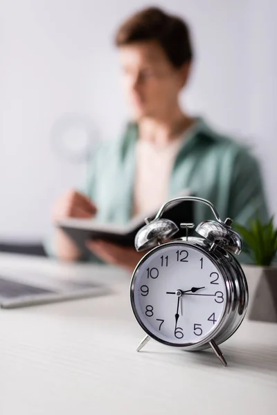 Selective focus of alarm clock on table and man holding notebook near laptop at home, concept of time management — Stock Photo