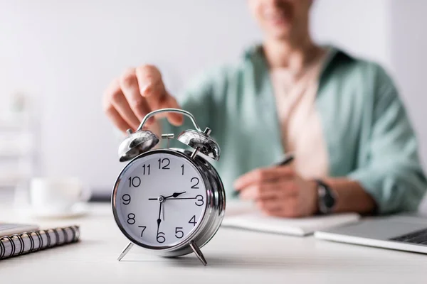 Selective focus of man pulling hand to alarm clock near laptop and notebooks on table, concept of time management — Stock Photo