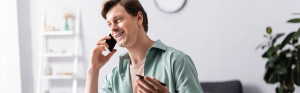 Панорамная ориентация позитивного человека, говорящего на смартфоне и держащего ручку дома, зарабатывающего онлайн концепцию — стоковое фото