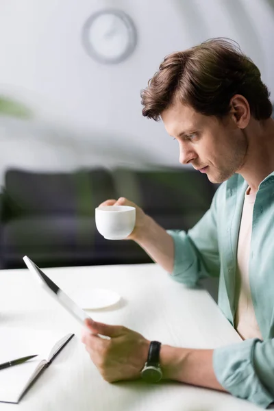 Selektiver Fokus eines gutaussehenden Mannes, der eine Tasse Kaffee hält und ein digitales Tablet am Tisch benutzt, Konzept des Online-Verdienens — Stockfoto