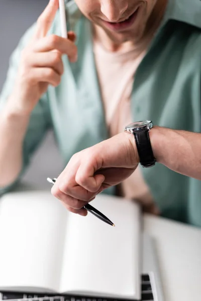 Cropped view of smiling freelancer checking time while talking on smartphone and holding pen, concept of time management — Stock Photo