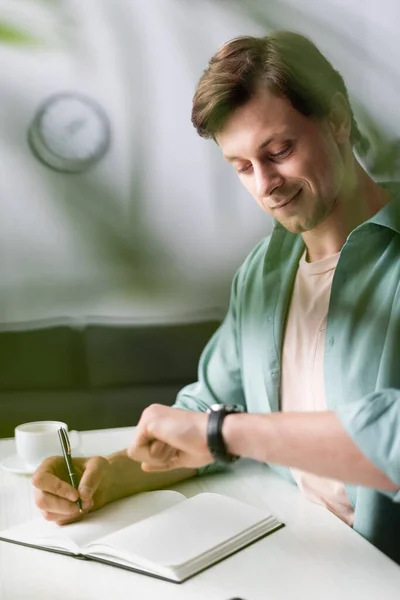 Selective focus of smiling freelancer checking time while writing on notebook near cup of coffee, concept of time management — Stock Photo