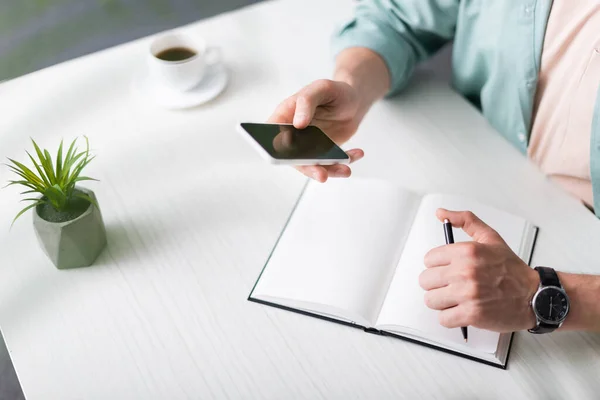 Vista ritagliata dell'uomo che tiene smartphone e penna vicino al notebook e alla tazza di caffè sul tavolo a casa, guadagnando concetto online — Foto stock