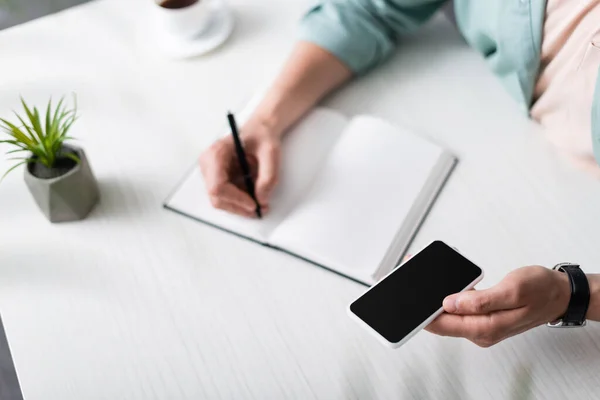 Ausgeschnittene Ansicht eines Mannes, der sein Smartphone mit leerem Bildschirm hält, während er am Tisch am Notizbuch schreibt und Online-Konzept verdient — Stockfoto