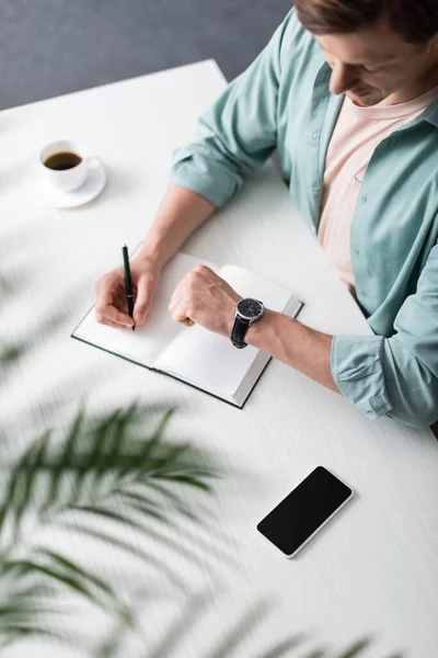 Selective focus of freelancer checking time while writing on notebook near smartphone and coffee on table, concept of time management — Stock Photo