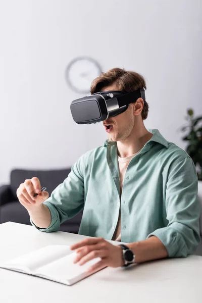 Selective focus of shocked man in vr headset holding pen near notebook on table — Stock Photo
