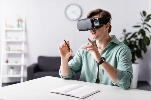 Sonriente hombre en vr auricular celebración de la pluma cerca de cuaderno abierto en la mesa — Stock Photo