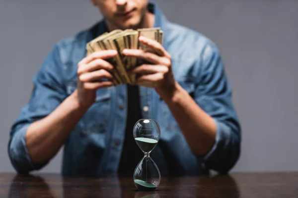 Foco seletivo de ampulheta na mesa e homem segurando dinheiro isolado em cinza, conceito de gerenciamento de tempo — Fotografia de Stock
